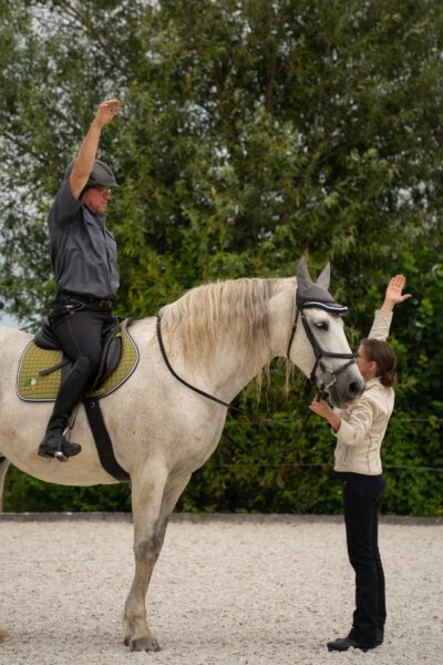 Reiter bei Uebung mit Schimmel Ein Mann mit Reithelm streckt auf einem Schimmel den Arm nach oben, eine Frau am Boden fuehrt das Pferd und hebt die Hand